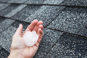 A hand holds a piece of hail the size of a golf ball against a black roof
