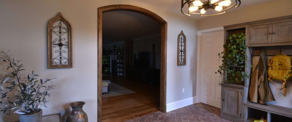 Finished residential front room home remodel at an angle with wood trim around an archway and a wooden coat rack against one wall. An industrial light fixture hangs from the ceiling