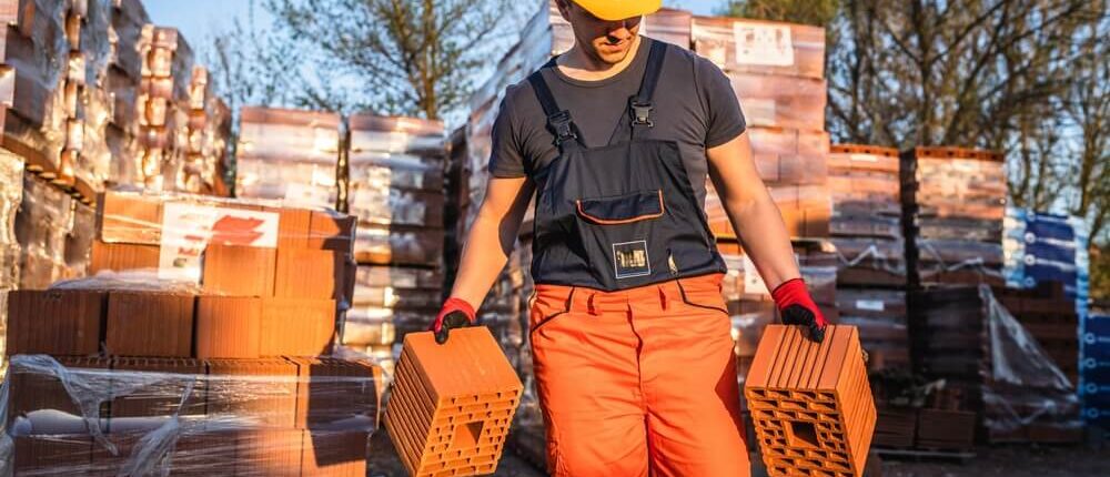 A construction worker in orange pants and a yellow hard hat carries building materials as he walks